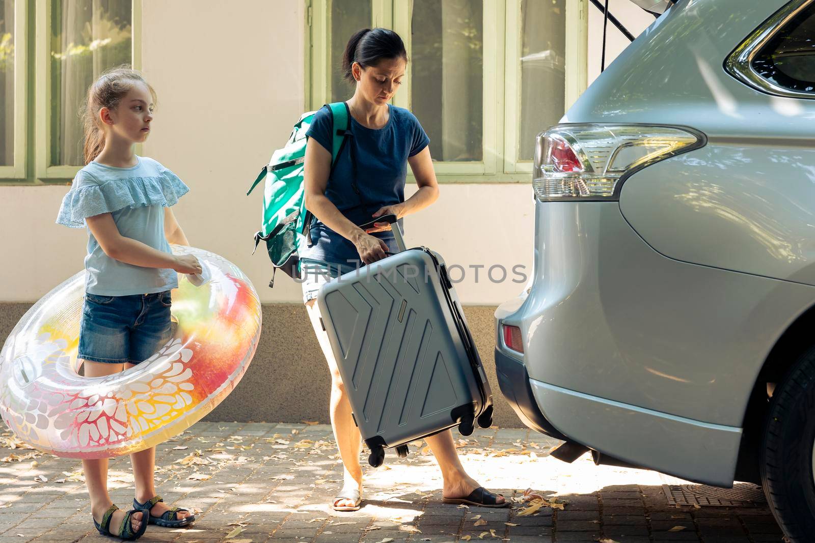 Mother and child travelling on vacation, loading baggage trolley and inflatable in vehicle trunk to go to seaside destination during summer holiday. Woman and little girl leaving on road trip.