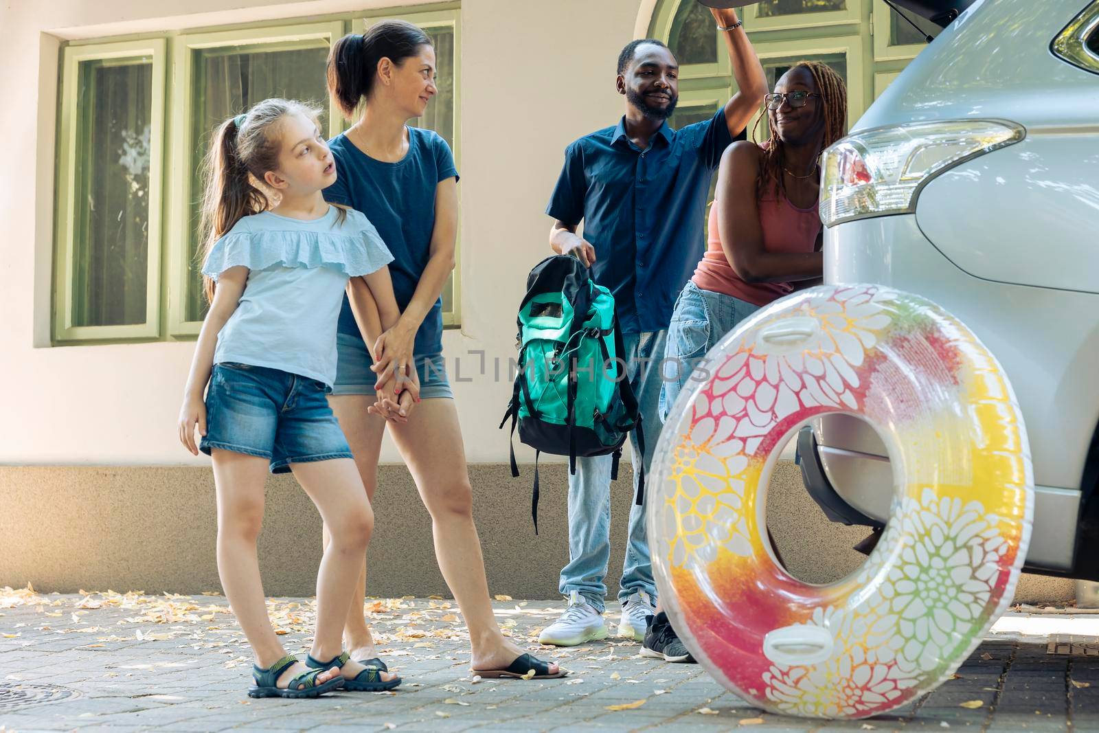 Mixed family and friends leaving on vacation trip, loading inflatable and trolley bags in trunk of vehicle to travel on summer holiday journey. Going to seaside destination with smiling people.