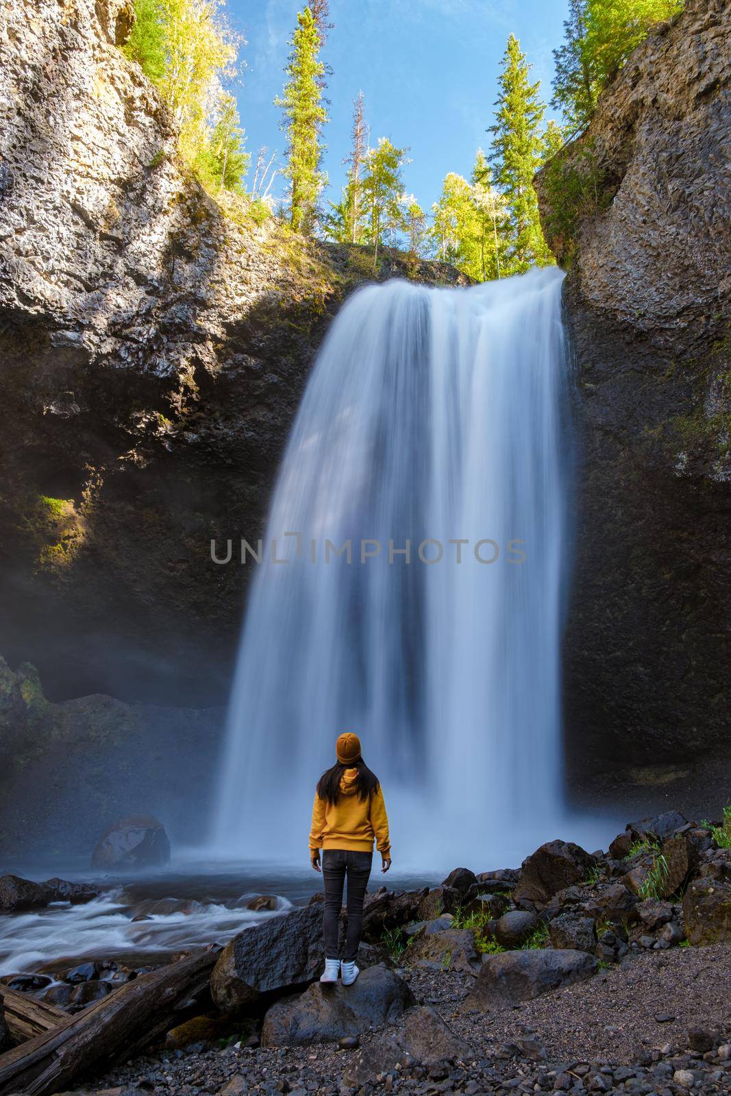 Moul waterfall in Canada, couple visit Moul Falls, the most famous waterfall in Wells Gray Provincial Park in British Columbia, Canada. Asian woman with a yellow sweater and hat looking at a waterfall