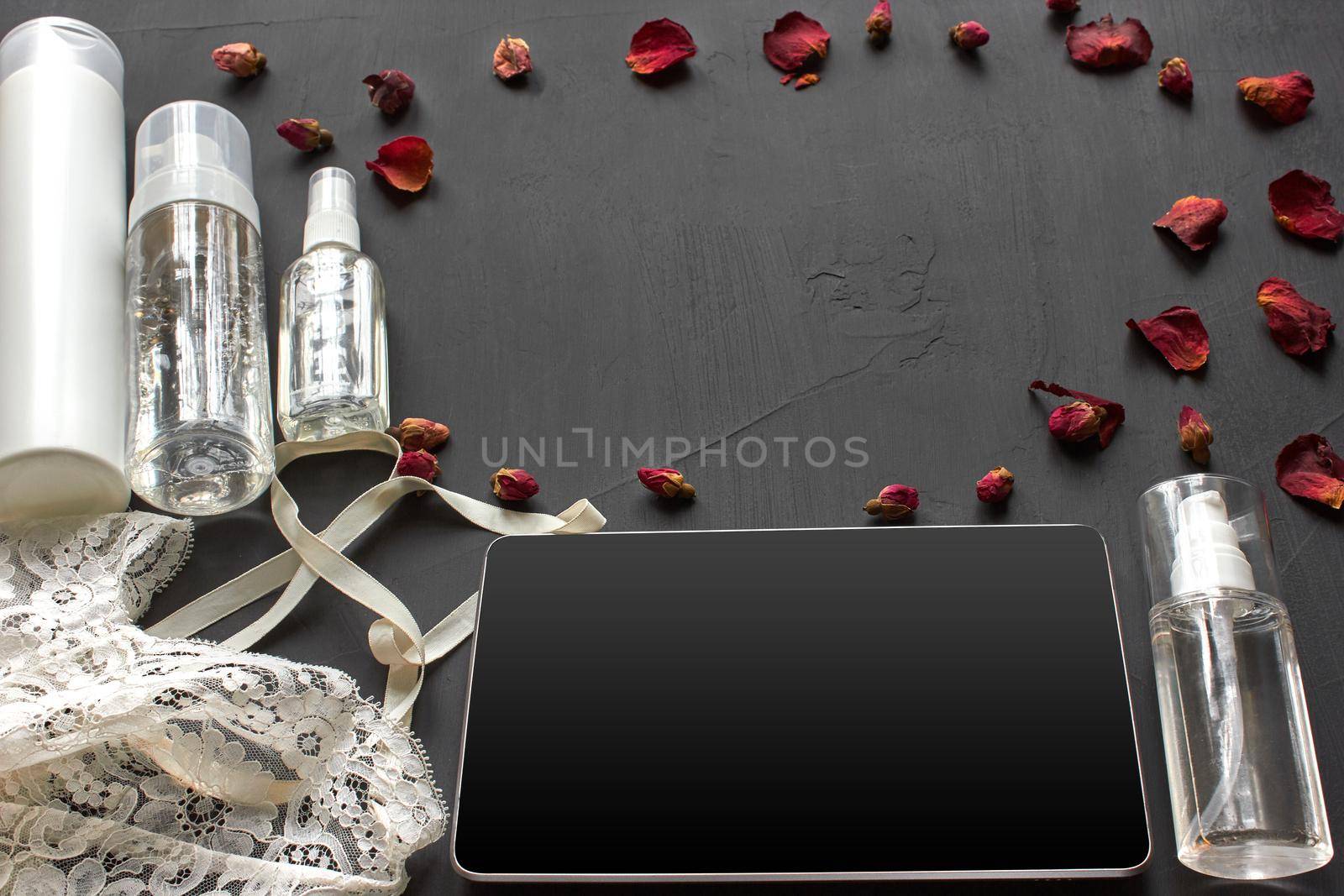 Skin care cosmetic products without label, white lace lingerie and a tablet on a black background with copy space, decorated with dried rose buds. Beauty trends, top view. Branding mockup.
