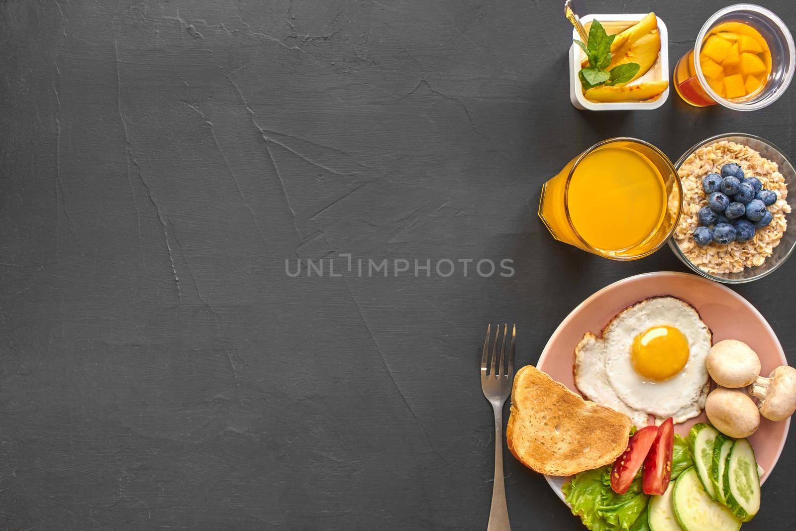 Top view of healthy and delicious breakfast on a black background with copy space. Fresh vegetables, scrambled eggs, toast are on a plate for tasty and nutritious meal. Oat flakes with a blueberry, yoghurt, orange juice and fork are nearby.