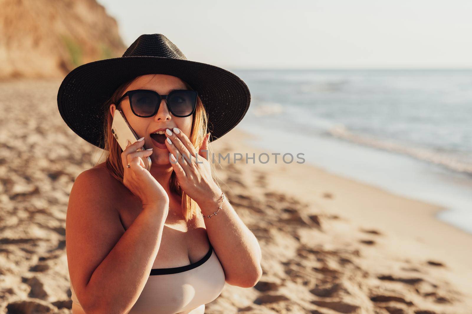 Young Woman Emotionally Expresses Surprise While Talking on the Phone on Sandy Beach by the Sea by Romvy