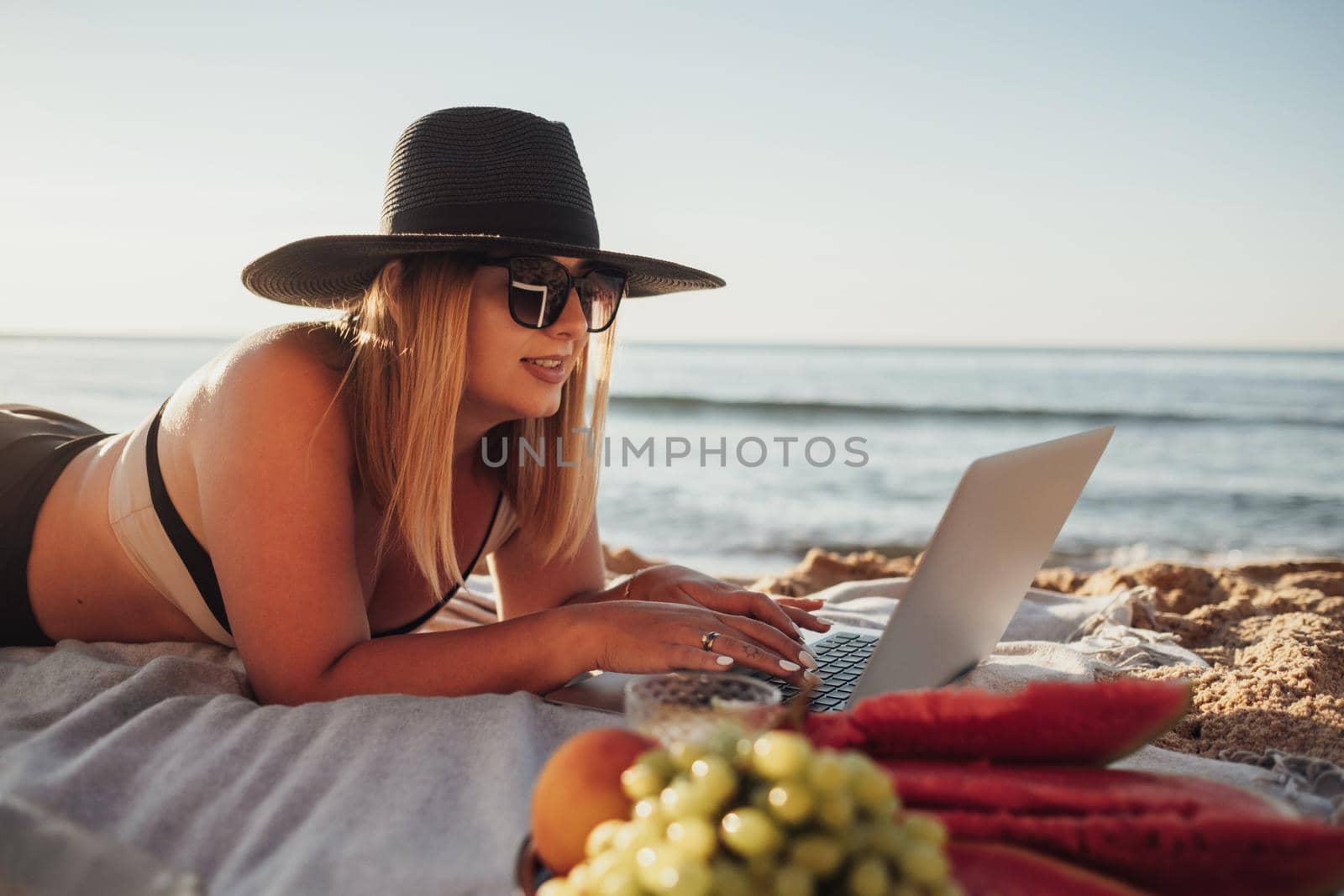 Pretty Young Woman in Sunglasses and Hat Using Laptop, Enjoying Picnic on Vacation While Lying on Sandy Beach by the Sea by Romvy