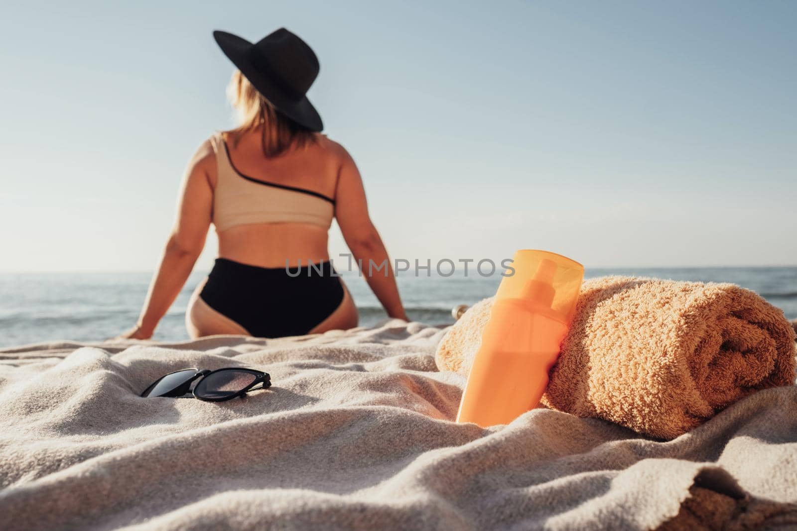 Vacation Time Concept, Sunscreen Lotion and Towel in Focus, Back View of Woman Sitting on the Beach by the Sea by Romvy