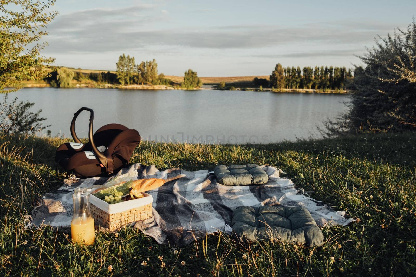 Picnic Set Outdoors by Lake at Sunset, Child Car Seat, Fruit Basket and Bottle of Juice on Plaid