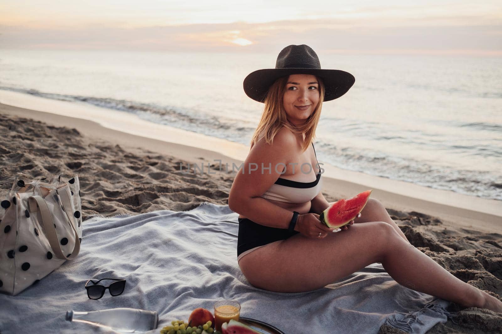 Young Pretty Woman Holding Slice of Watermelon and Looking Away While Sitting on Sandy Beach by the Sea at Sunrise by Romvy