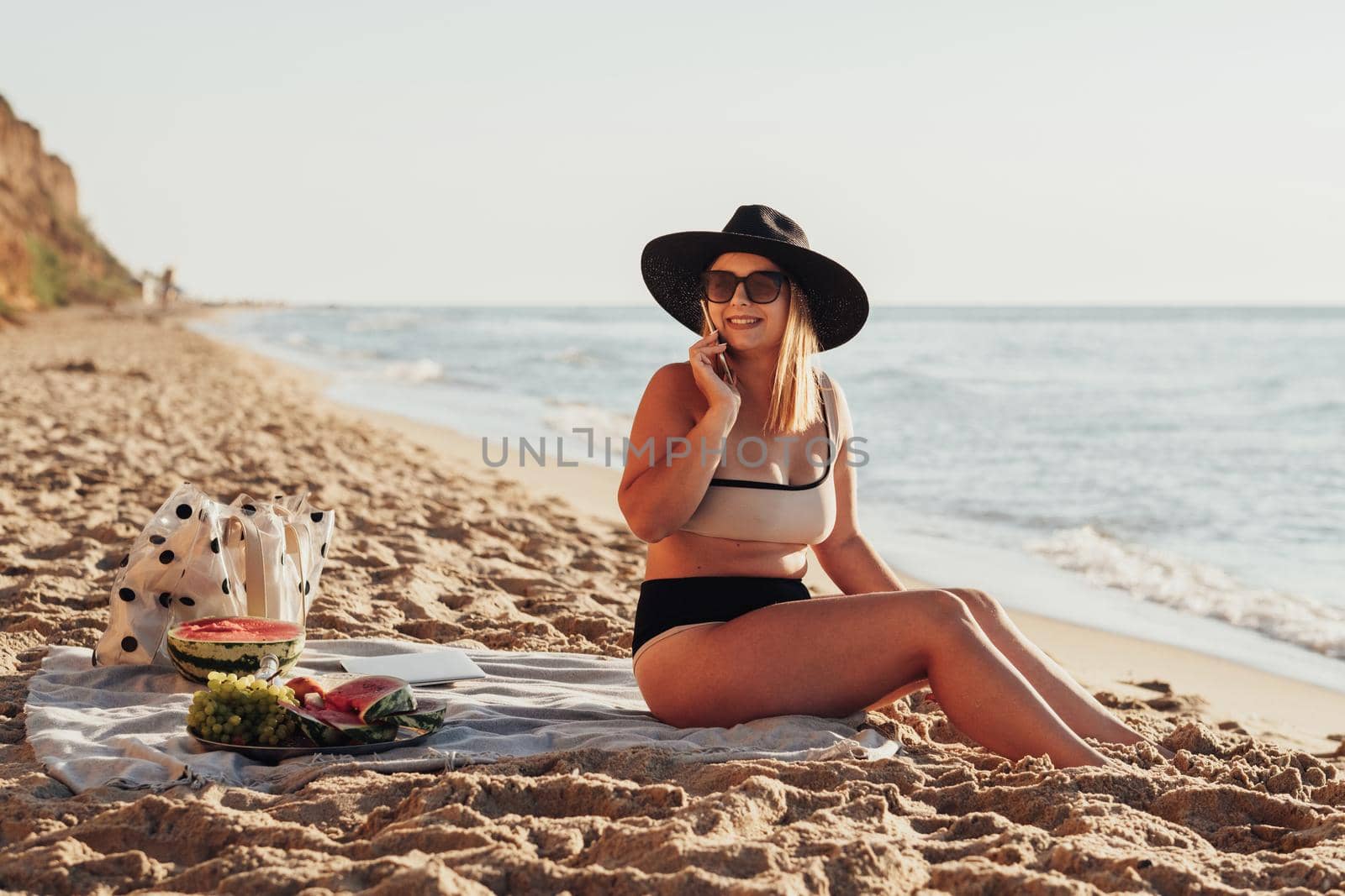 Smiling Young Woman Looking Away and Talking on the Phone While Sitting on Sandy Beach by the Sea by Romvy