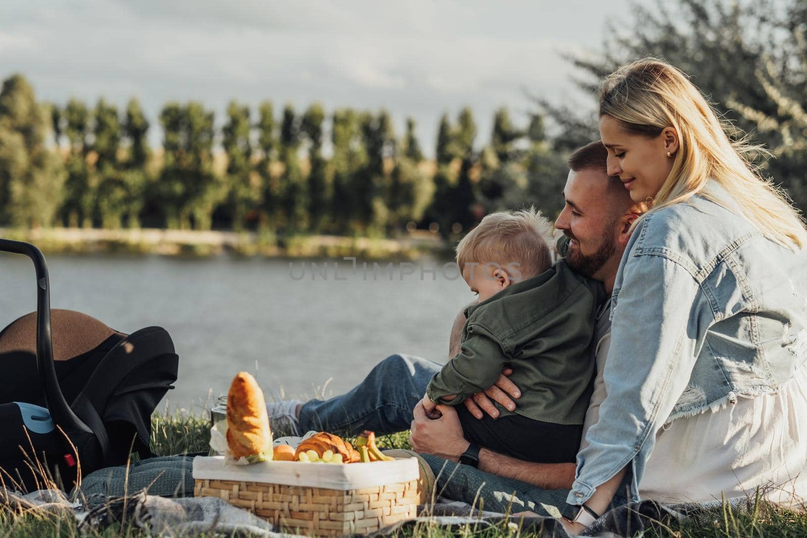 Happy Young Family, Mother and Father with Their Little Son Having Picnic Outdoors by Lake at Sunset