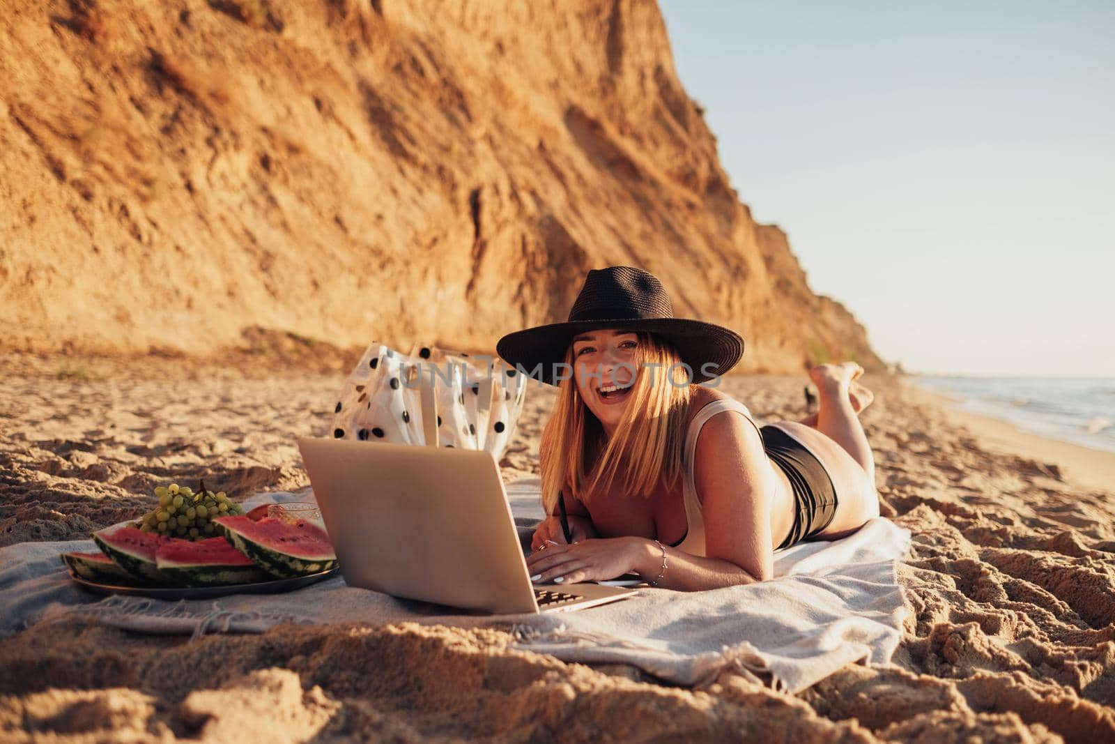 Cheerful Young Woman in Swimsuit and Hat Smiling Into the Camera While Working on Laptop on Vacation by Sea