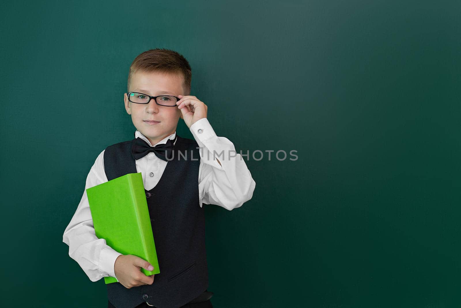 Happy cute clever boy in glasses with book in his hand. Child is ready to answer with a blackboard on a background. First time to school. Back to school.