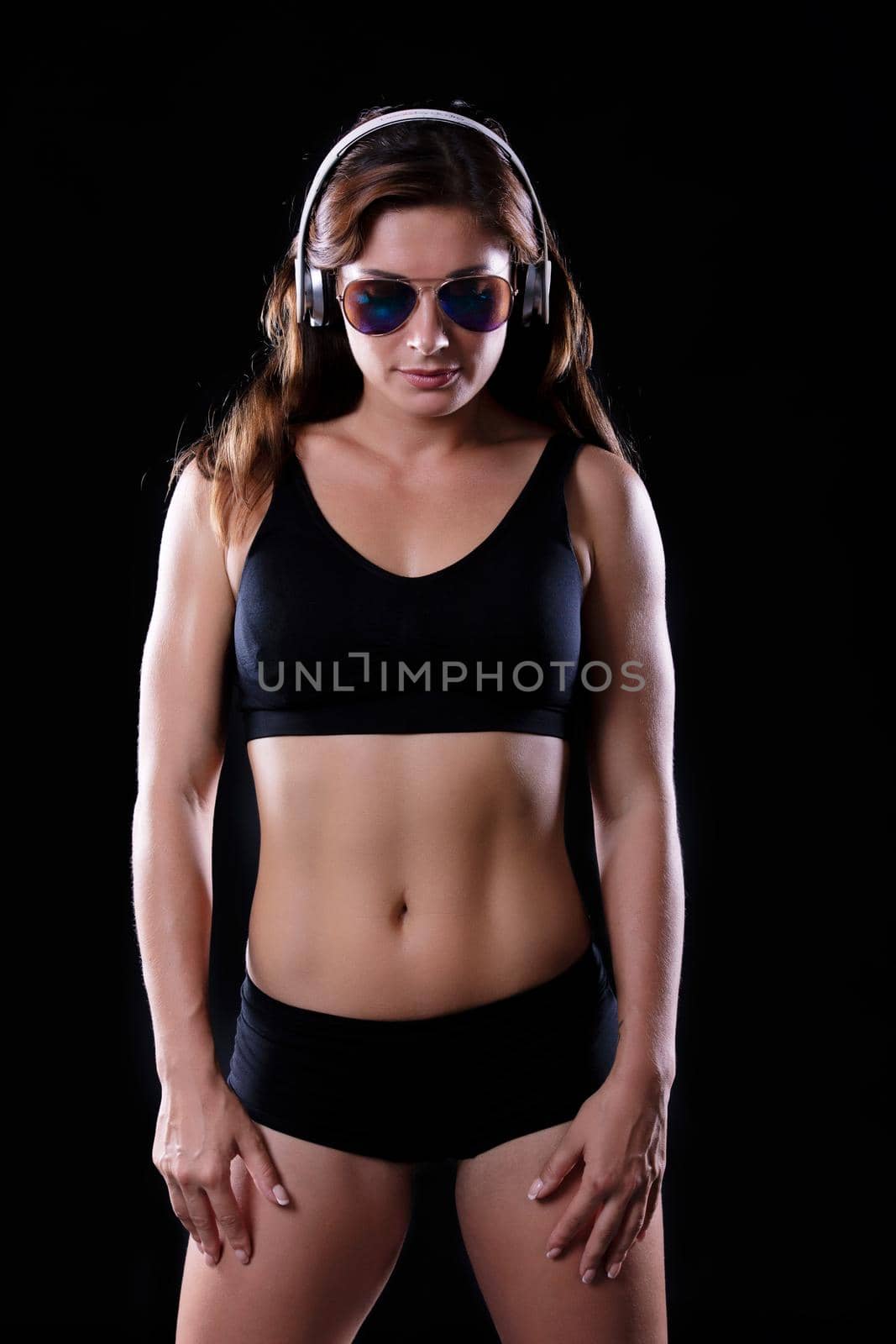 Sporty female brunette girl showing off her perfect body on black background with headphones and sunglasses