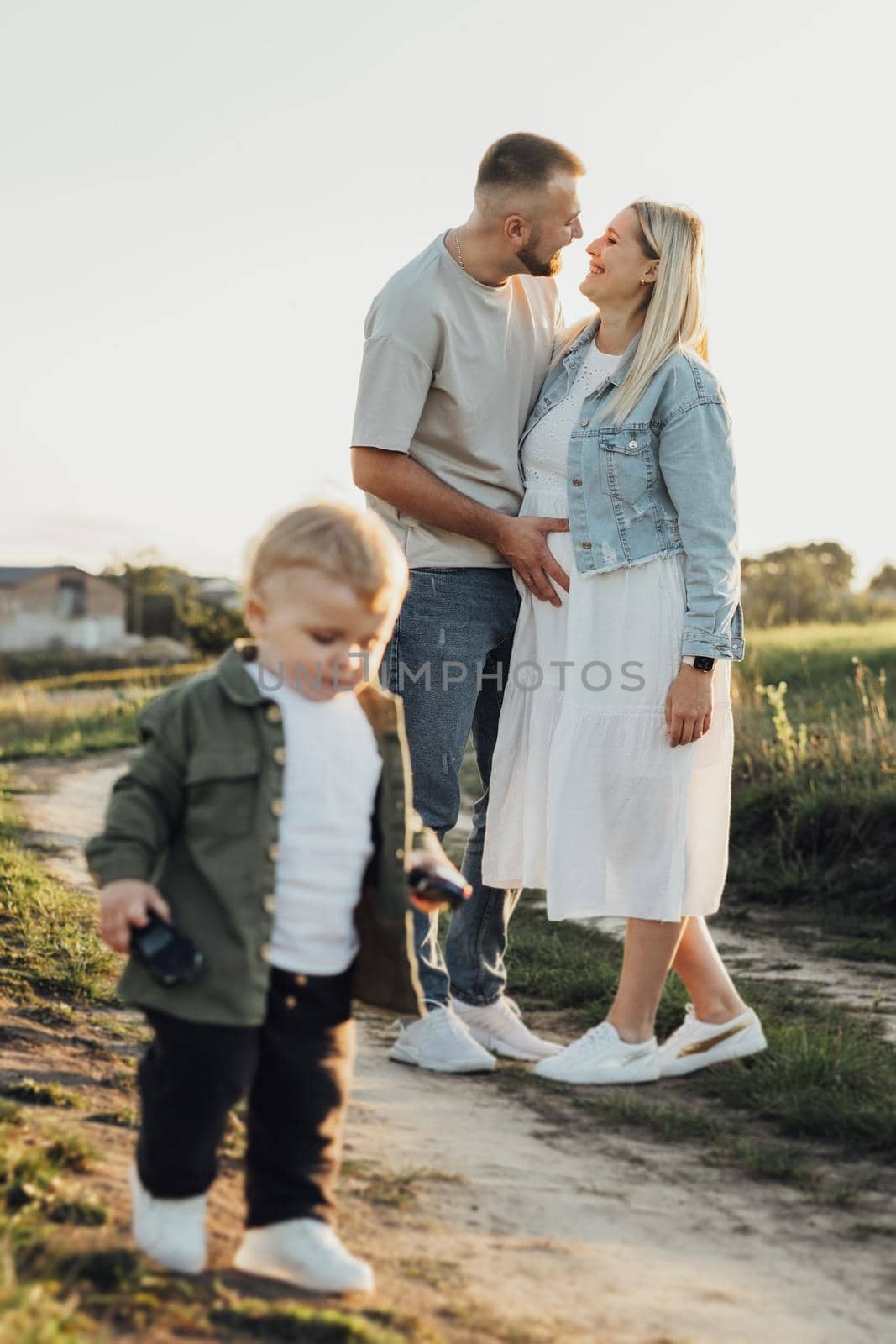 Happy Young Family Outdoors, Father Holding His Pregnant Wife by Belly While Their Little Son Playing Around Them