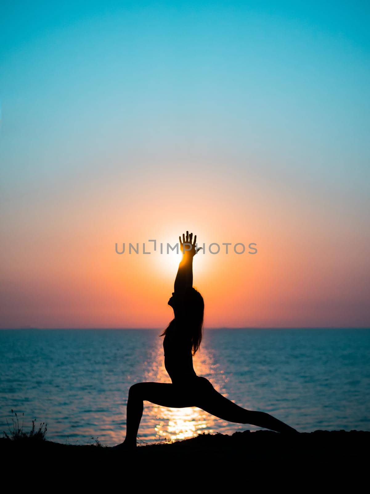 Young slim girl practicing yoga on mountain against ocean or sea at sunrise time. Silhouette of woman in rays of awesome sunset.