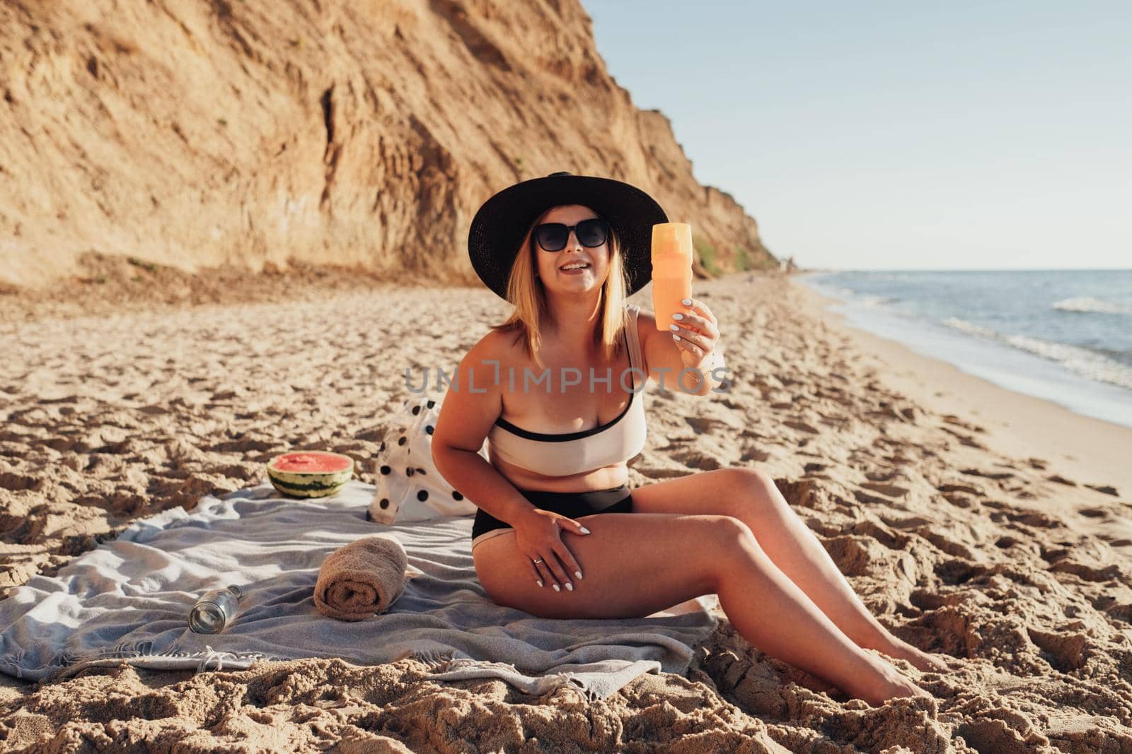 Young Woman Enjoying Vacation by the Sea, Holding Sunscreen Lotion While Sitting on Beach