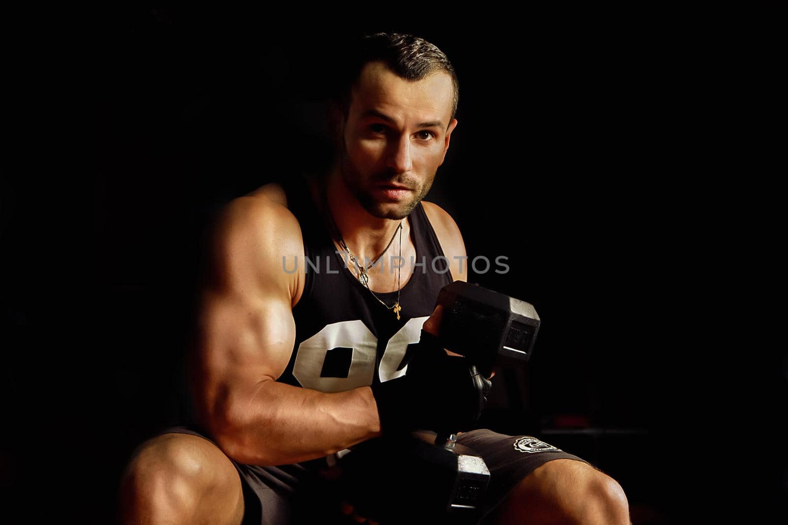 Close up portrait of a fit young man working out with steel dumbbells and looking at the camera on dark background