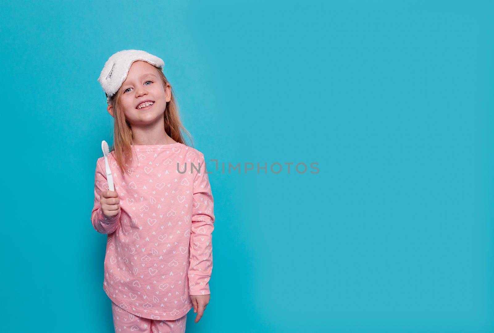 close-up of a blonde, caucasian girl in pink pajamas, a unicorn-like sleep mask. The child shows an toothbrush in the hand. Blue background, copy space. The concept of children's health, good sleep