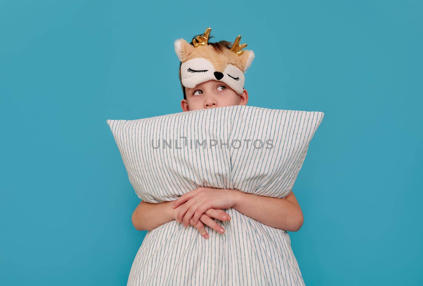 the cunning boy does not want to sleep, hugs the pillow and looks away. Child on a blue background with copy space. The concept of a naughty child, parenting, insomnia, health