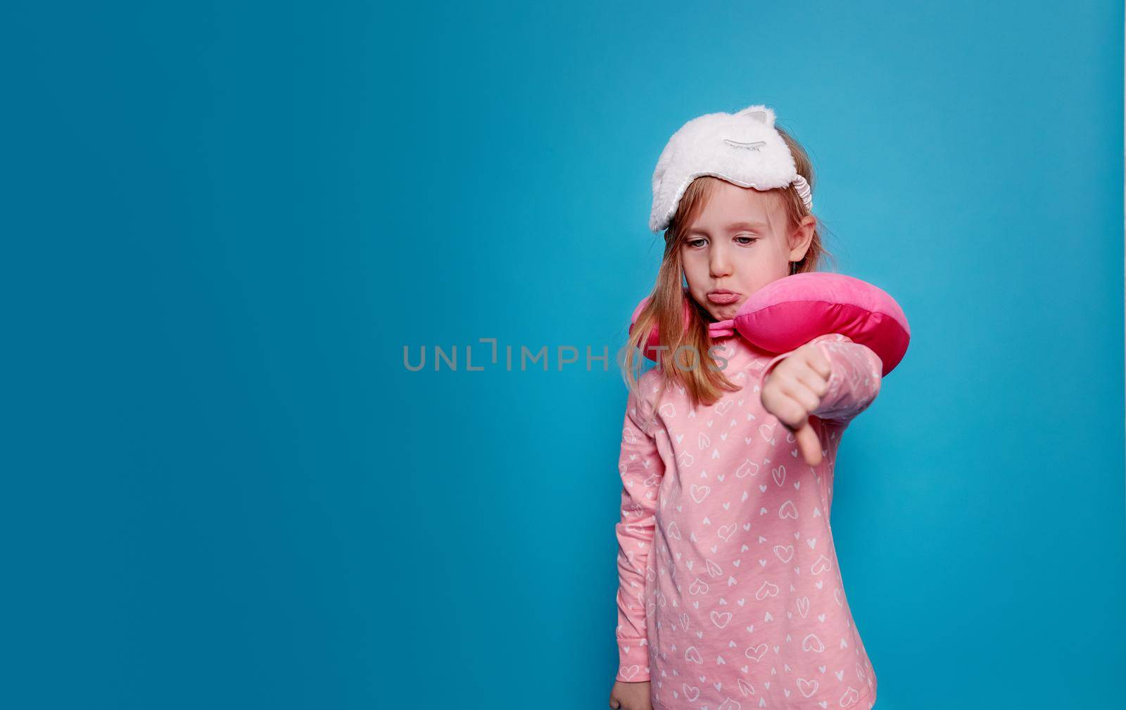 Sad girl, blonde, with a sleep mask in pink pajamas , a travel pink pillow around her neck. Blue background, copy space. The concept of insomnia, poor sleep, unhealthy. Bad linens. orthopedic pillow