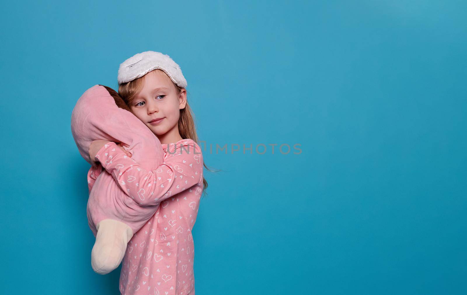 little girl in pajamas and sleep mask hugs toy on a blue background, copy space by Ramanouskaya