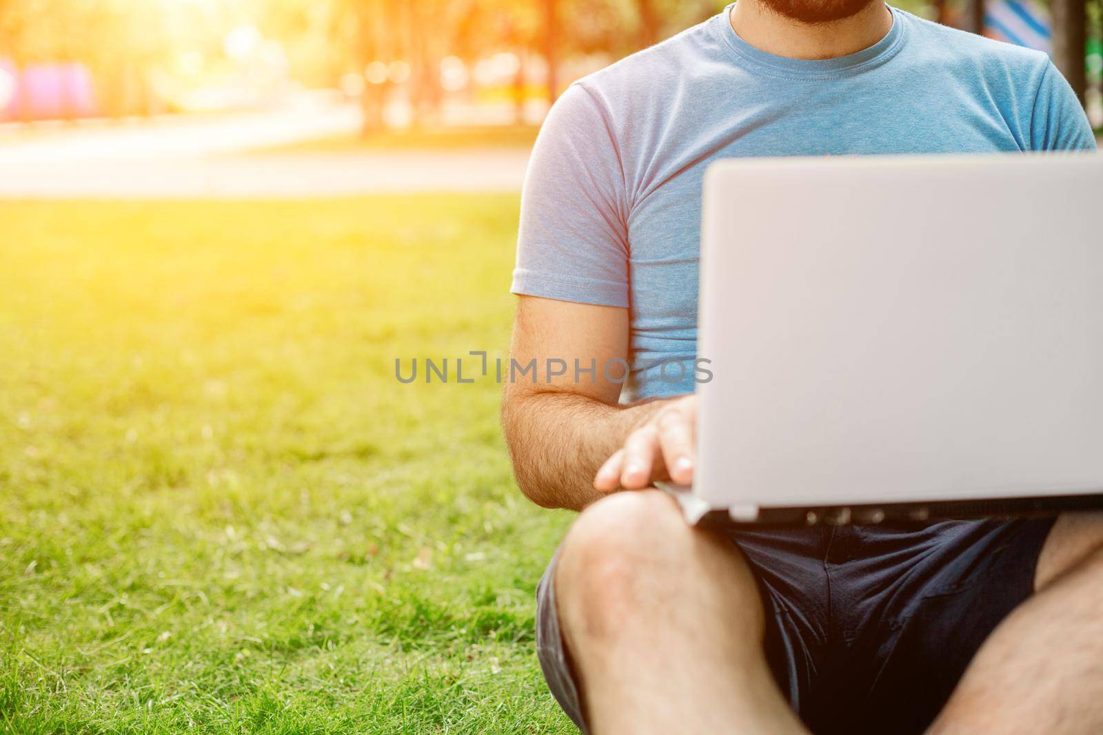 Young man using and typing laptop computer in summer grass. Freelancer working in outdoor park. Sun flare