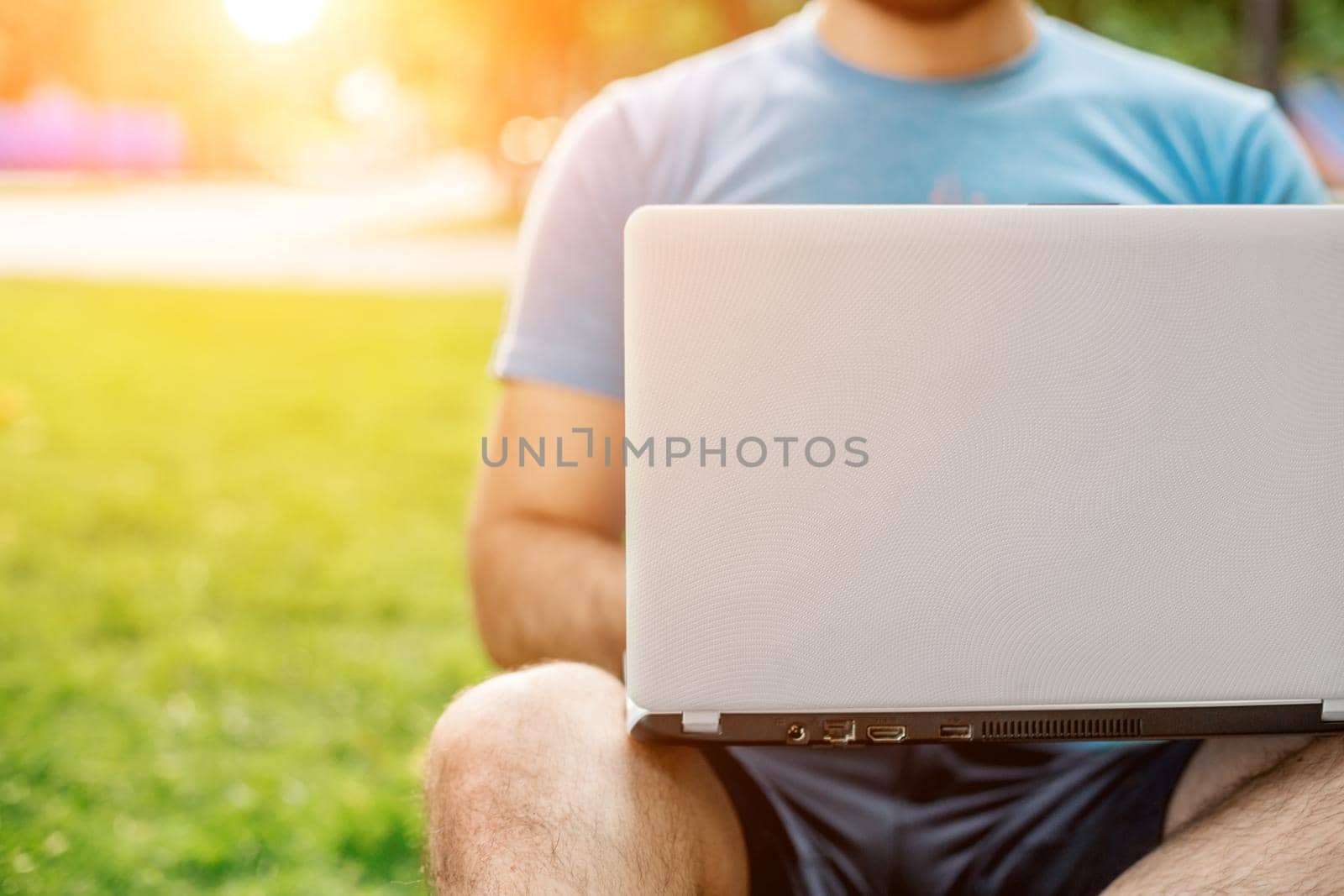 Young man using and typing laptop computer in summer grass. Freelancer working in outdoor park. Sun flare