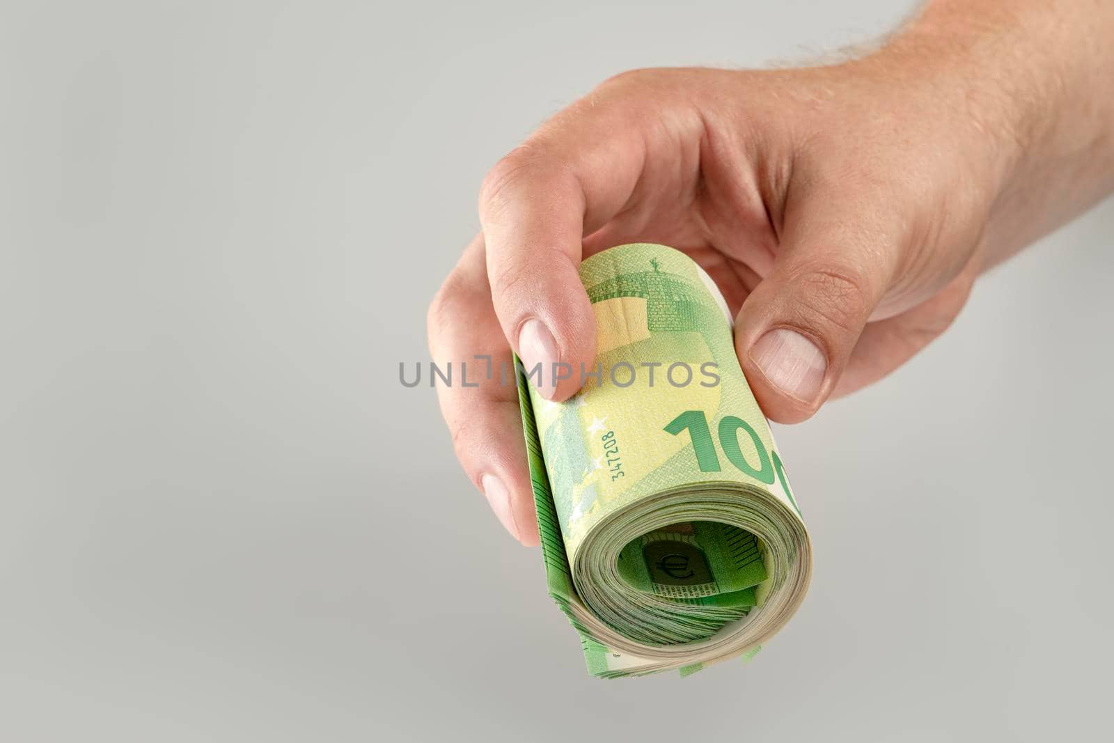 The hand holds a roll of 100 euro banknotes. Euro banknotes rolled up in a white man's hand on a gray background. The concept of financial assistance, real estate purchase, loan or insurance payment