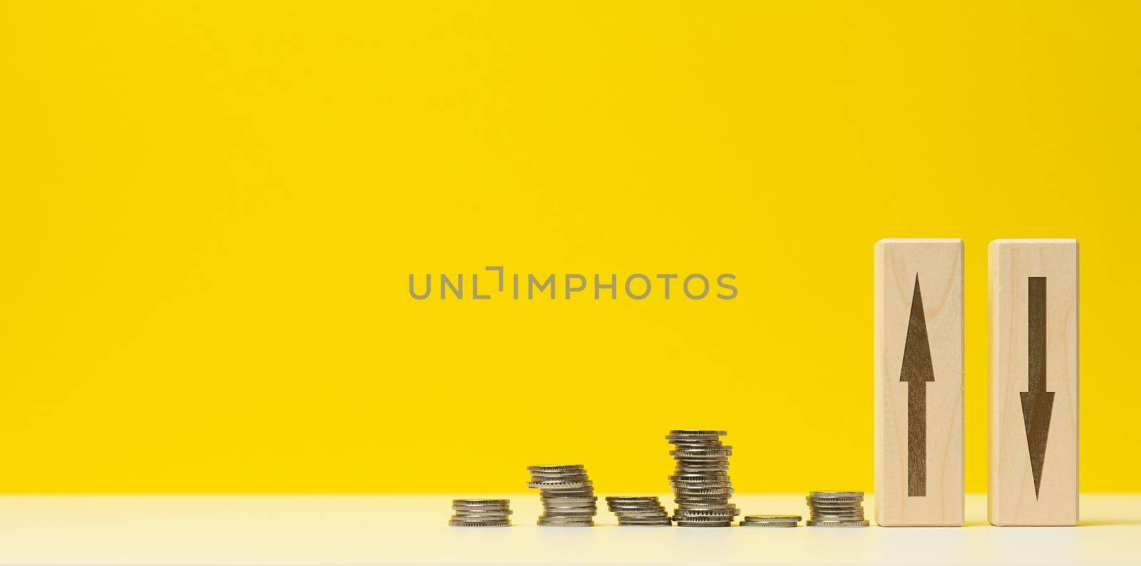 stacks of metal money and two wooden bars with arrows on a white table, yellow background. The concept of depreciation and depreciation of the exchange rate, the fall of the economy by ndanko