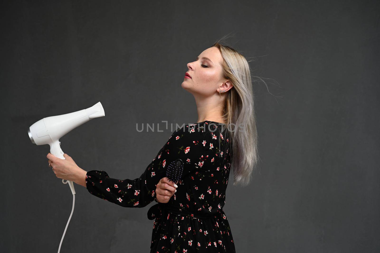 curly blondy woman using hair dryer on gray background. Making perfect curls by chichaevstudio