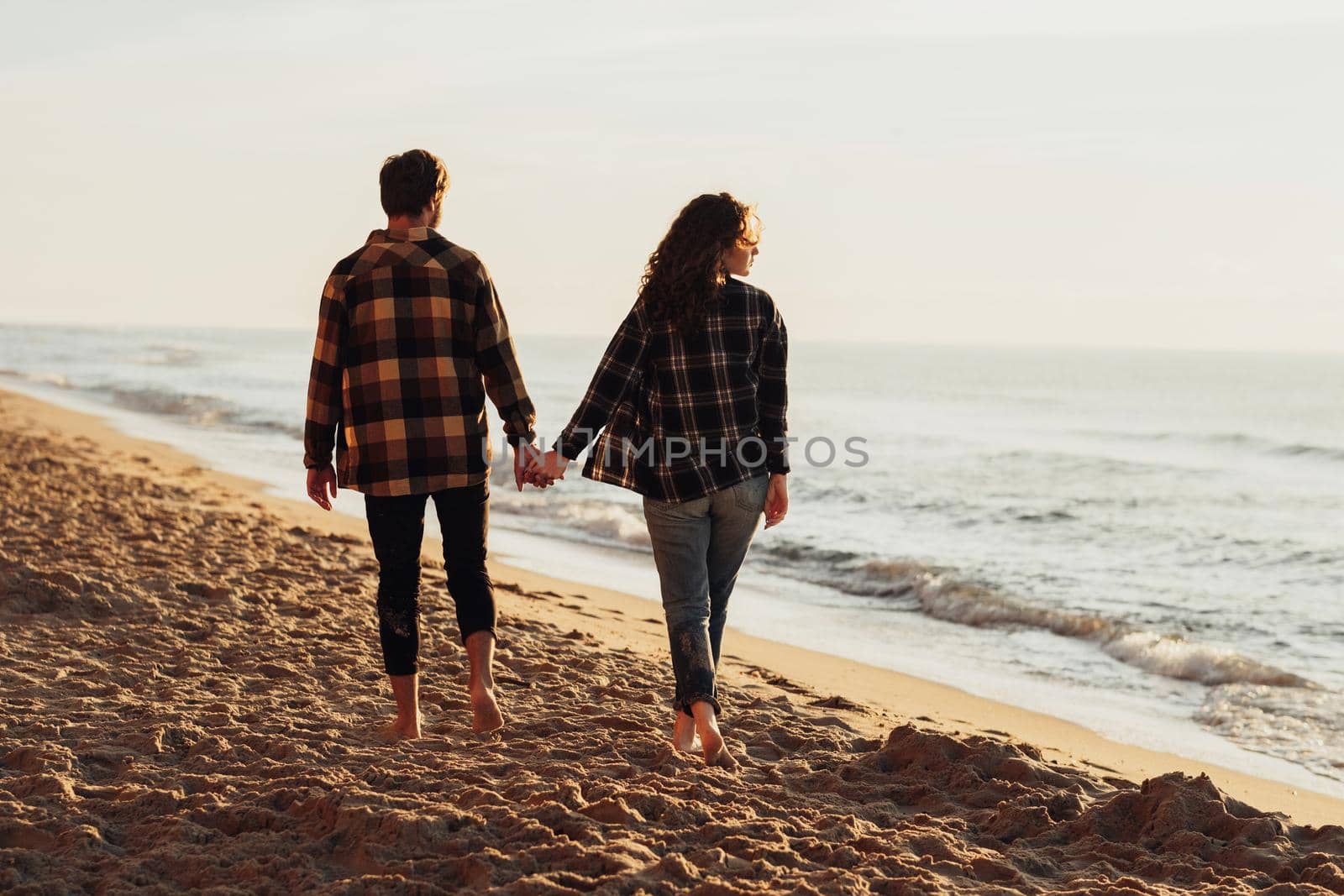 Young couple, woman and man holding hands together and walking along sea coast at dawn