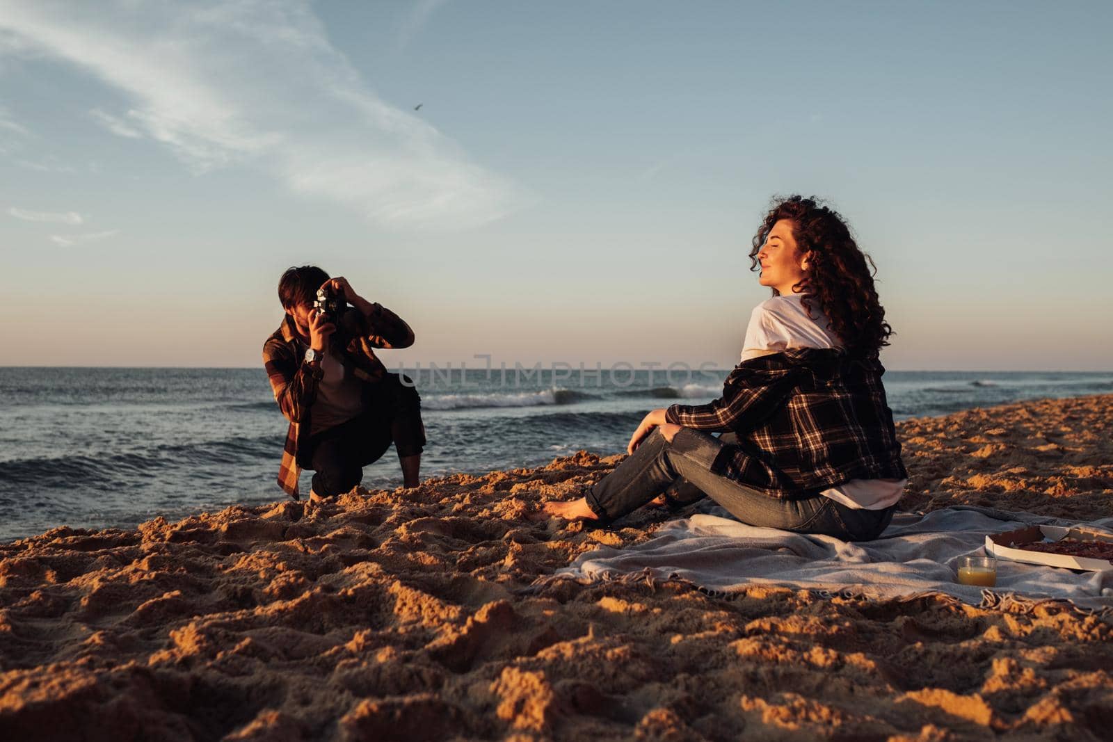Young man photography his curly haired girlfriend that sitting on the seashore at dawn, couple enjoying sunrise on the beach by Romvy