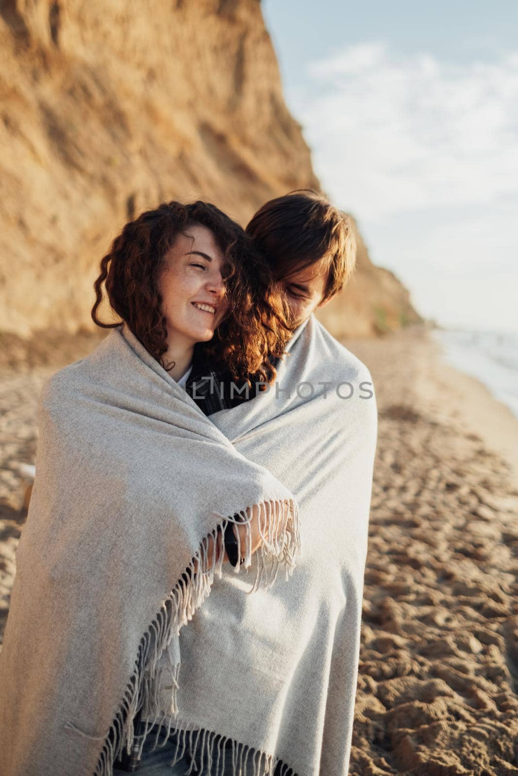 Young cheerful woman and man standing on the seashore wrapped in plaid at sunrise