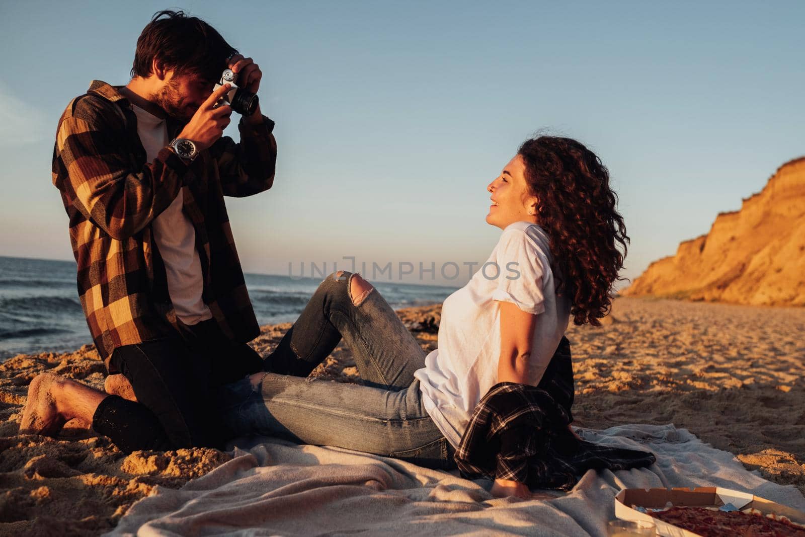 Young man photography his curly haired girlfriend that sitting on the seashore at dawn, couple enjoying sunrise on the beach by Romvy