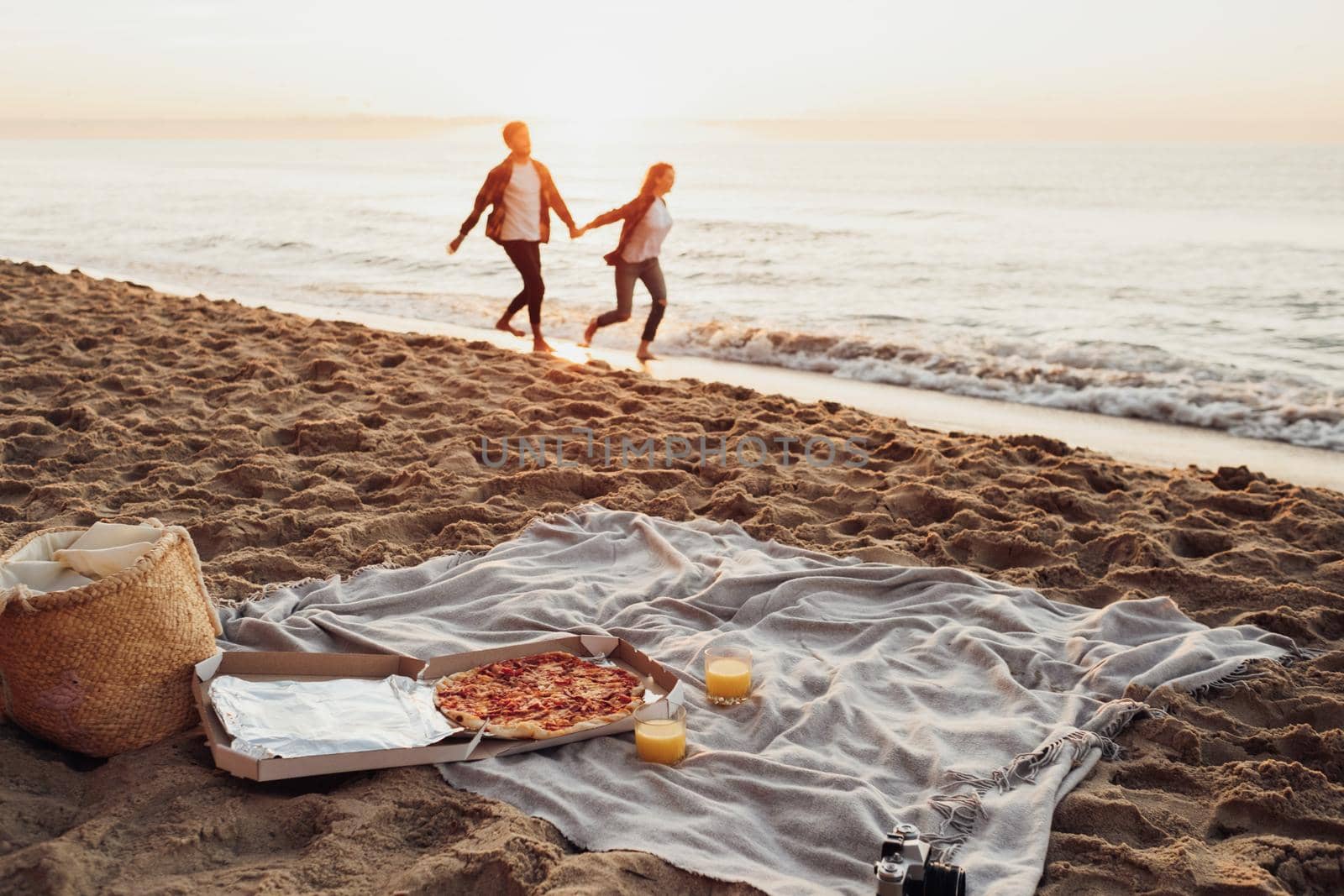 Focus on picnic set with pizza and juice, happy couple, woman and man holding by hands and running along the coastline together on background