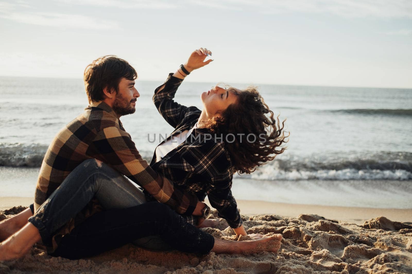 Young adult couple fall in love, curly haired woman sitting on man and enjoying of being together on seashore at sunrise