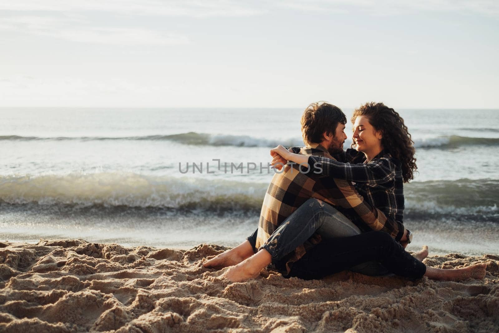 Young couple, curly haired woman with brunette man fall in love sitting and hugging on the seashore at sunrise by Romvy