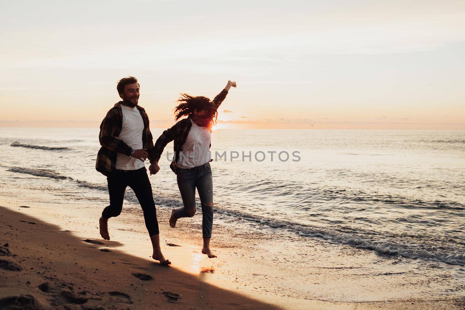 Happy young couple holding by hands and running along the coastline, woman and man meeting sunrise on sea together