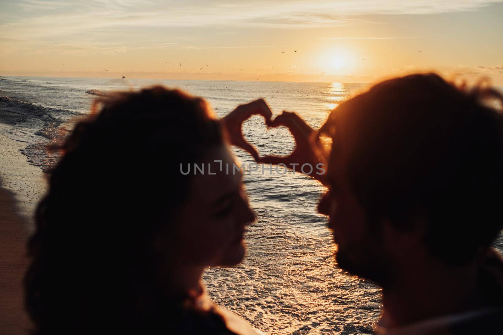 Man and woman showing heart by fingers against background of scenic sunrise, young couple meeting dawn on the seashore