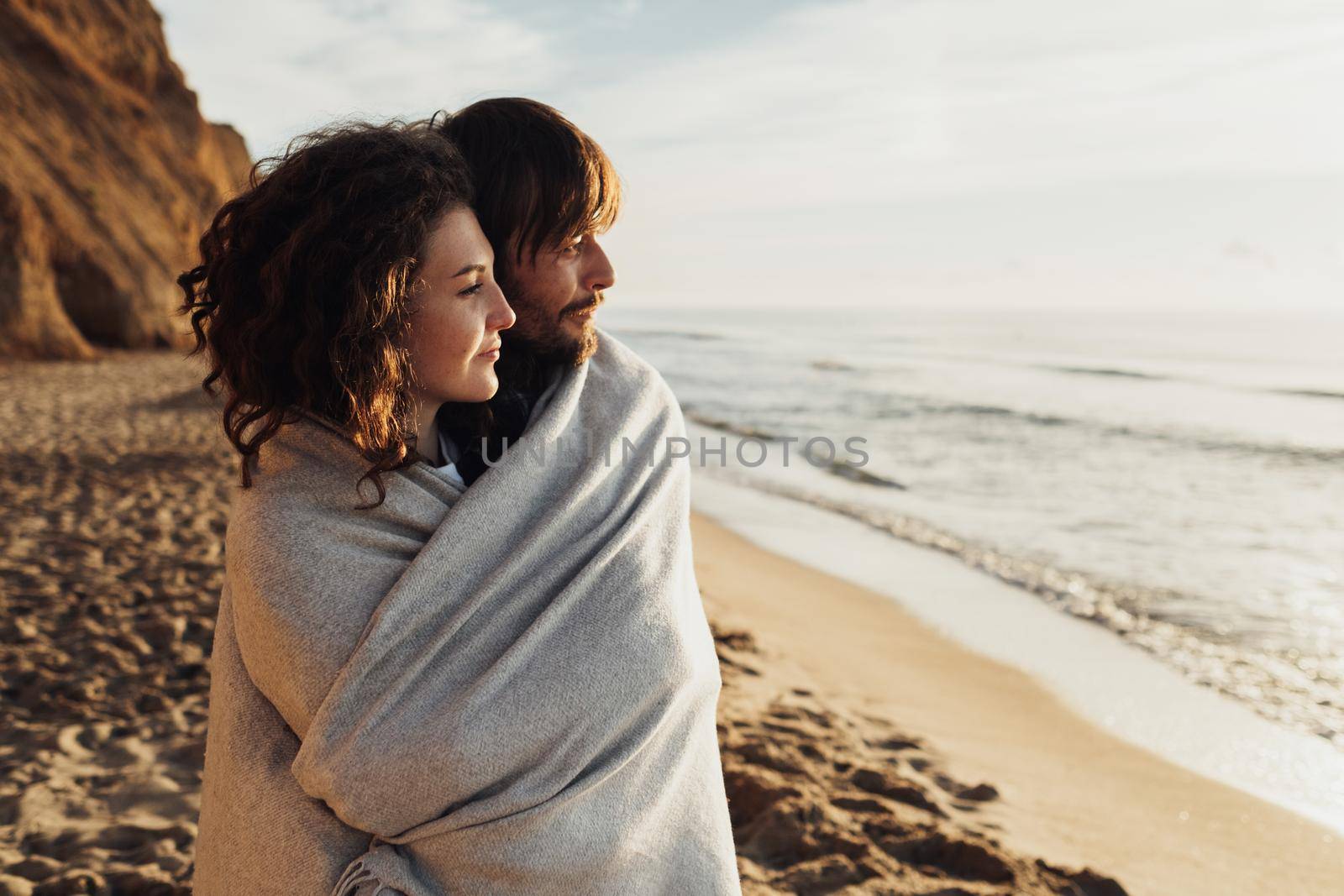 Young adult couple standing on the seashore wrapped in plaid and looking on sea waves at sunrise