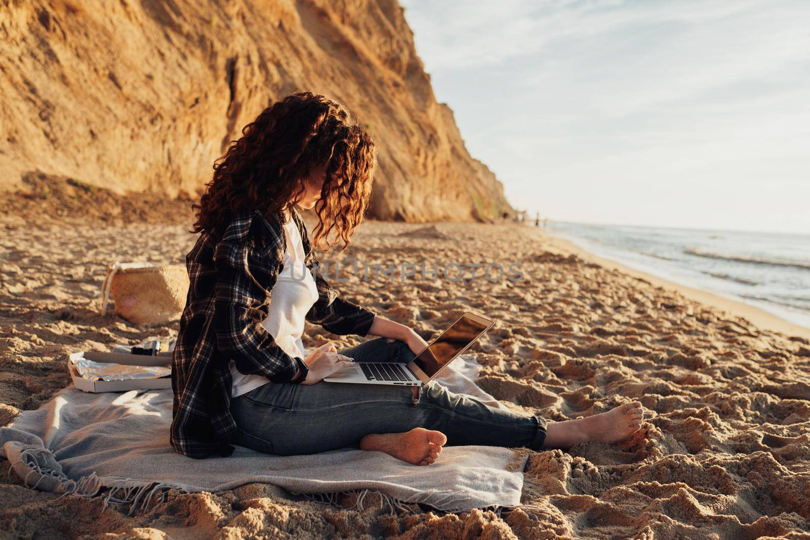 Curly haired woman working on laptop while sitting on the seashore at dawn with beautiful coastline on background