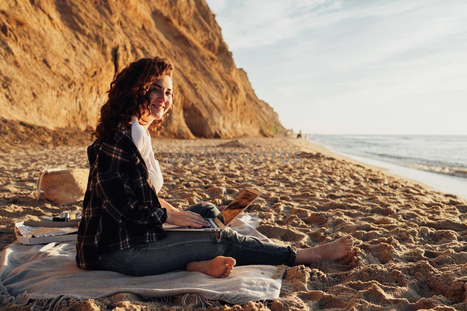 Curly haired woman working on laptop while sitting on the seashore at dawn with beautiful coastline on background
