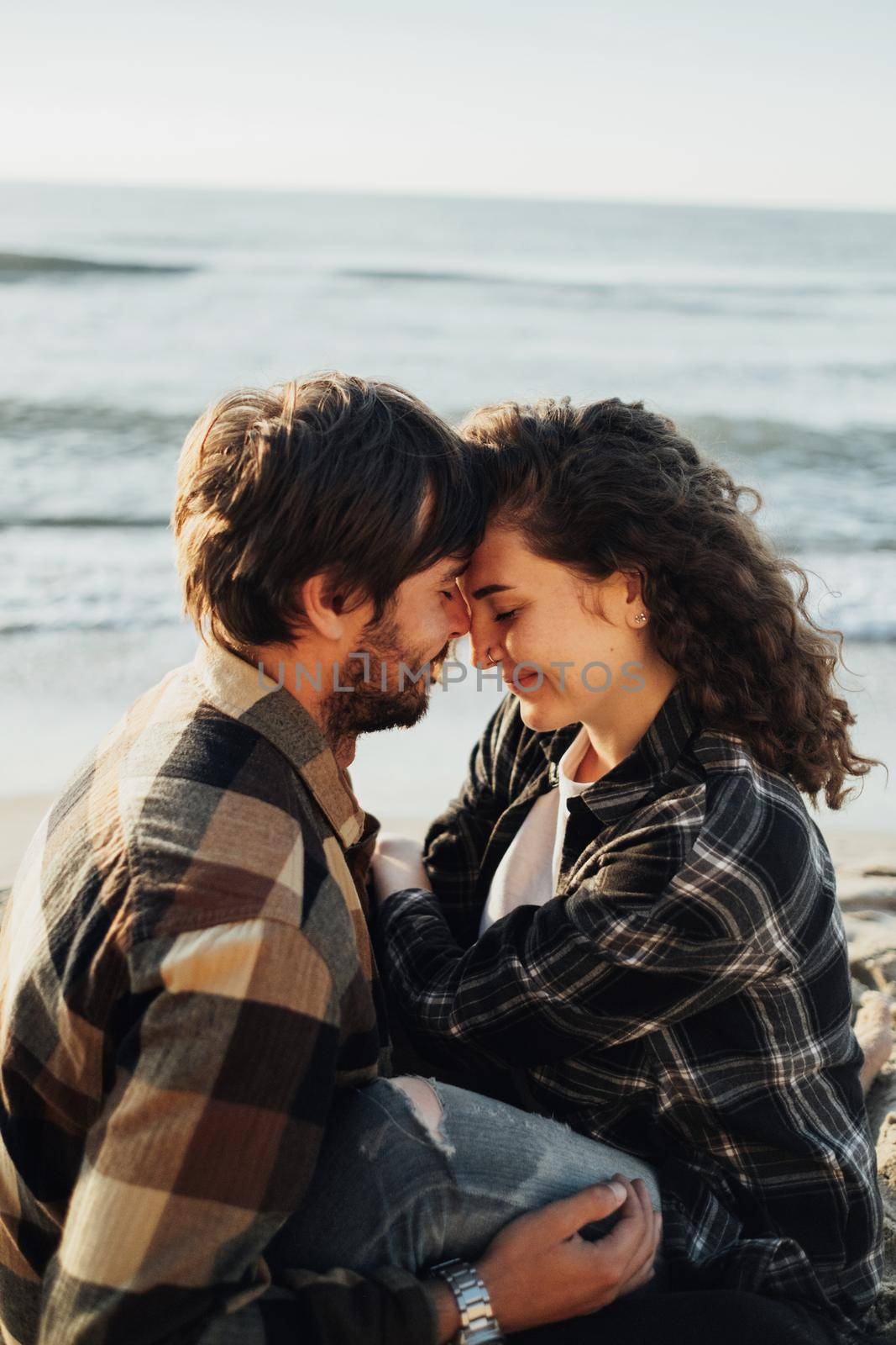 Close up of young woman and man hugging while sitting together on seashore with sea waves on the background at sunrise