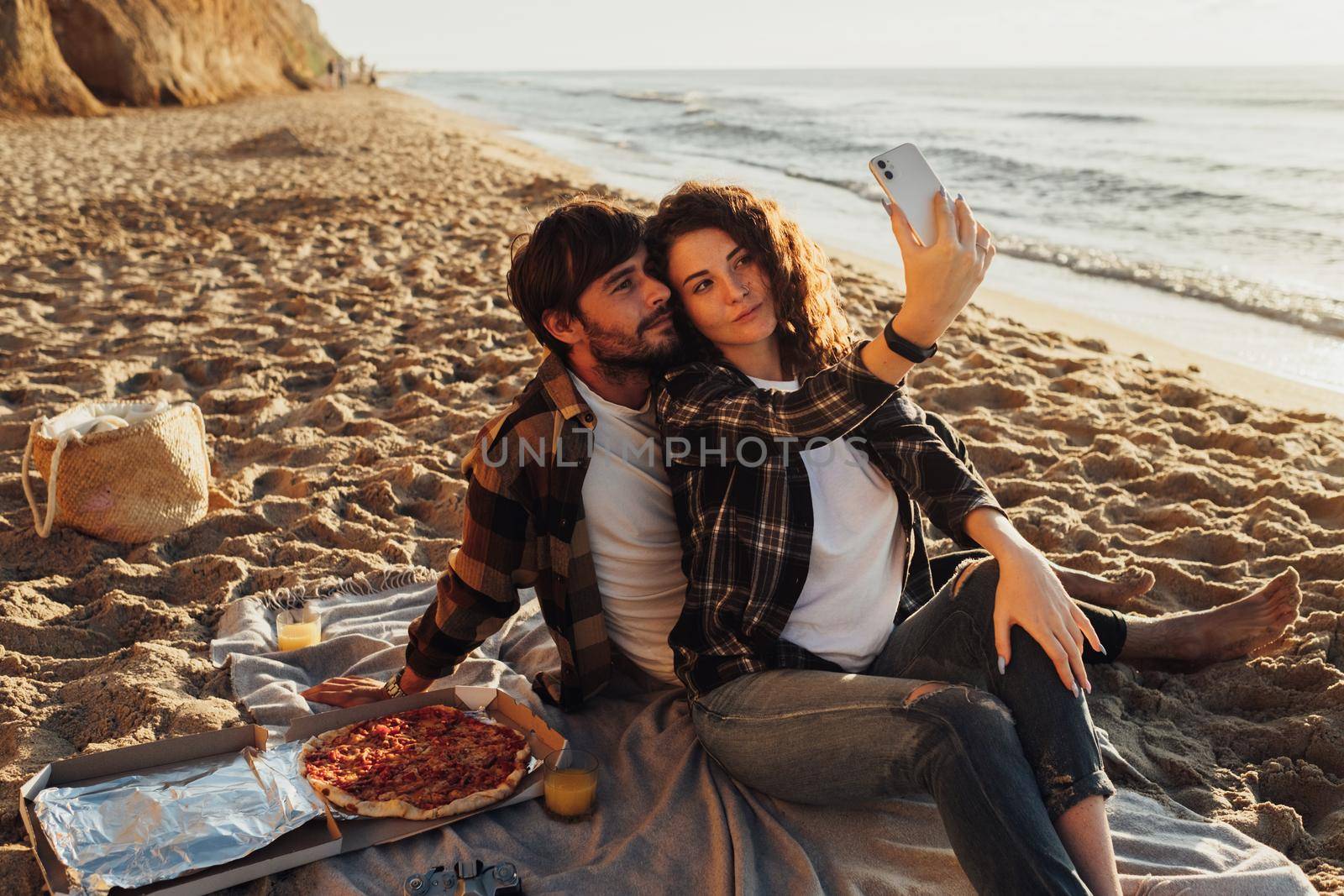 Happy young adult couple, woman and man making selfie together while enjoying picnic on the seashore at dawn by Romvy