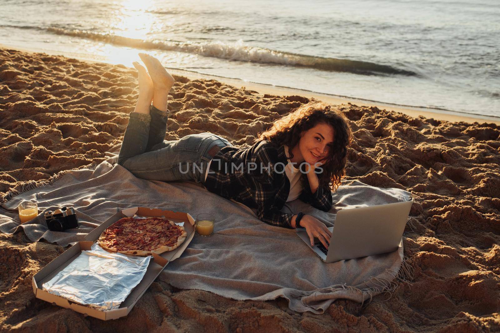 Happy curly haired woman eating pizza and working on laptop while laying on seashore at dawn