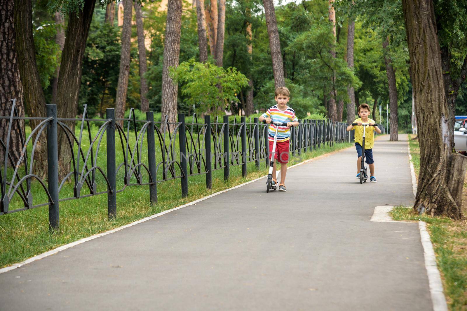 Two attractive European boys brothers, wearing red and white checkered shirts, standing on scooters in the park. They laughing, smiling, hugging and having fun. Active leisure time with kids.