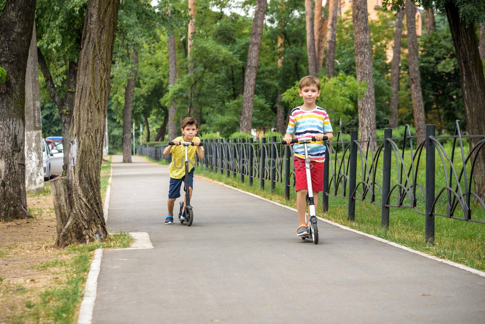 Two attractive European boys brothers, wearing red and white checkered shirts, standing on scooters in the park. They laughing, smiling, hugging and having fun. Active leisure time with kids.