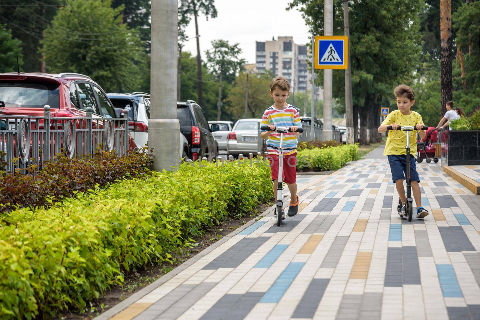 Two attractive European boys brothers, wearing red and white checkered shirts, standing on scooters in the park. They laughing, smiling, hugging and having fun. Active leisure time with kids.