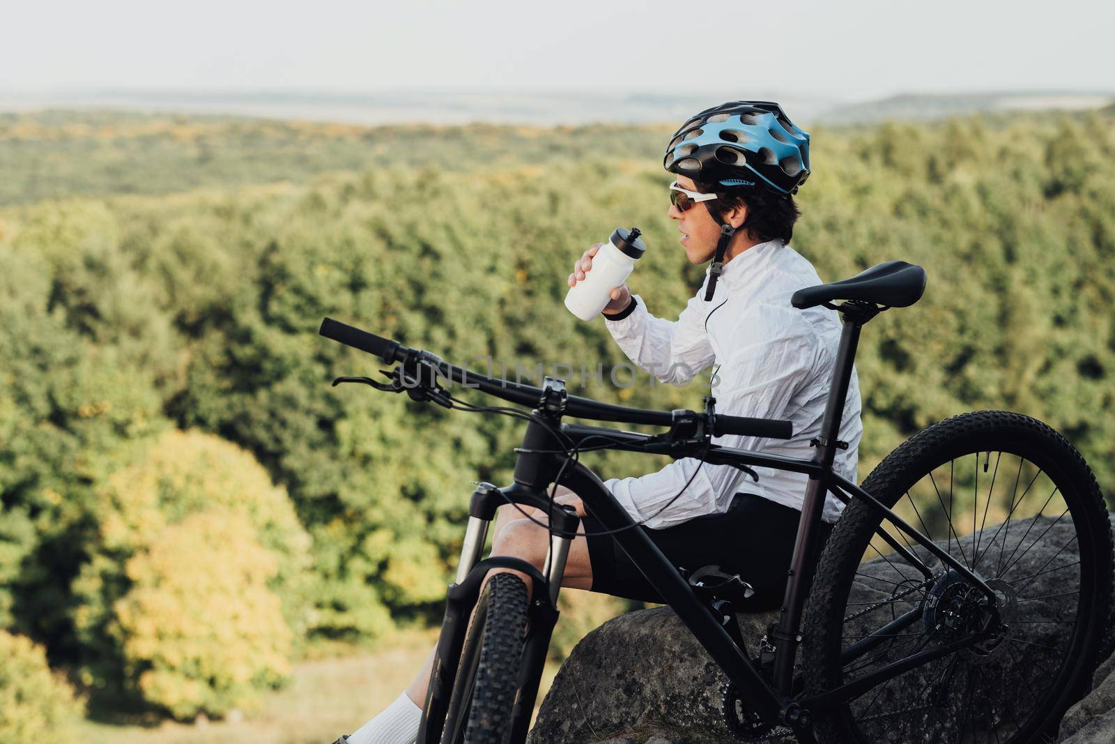Professional Male Cyclist Drinking Water from Bottle, Man Sitting Near His Bicycle During His Journey Outdoors in Countryside by Romvy