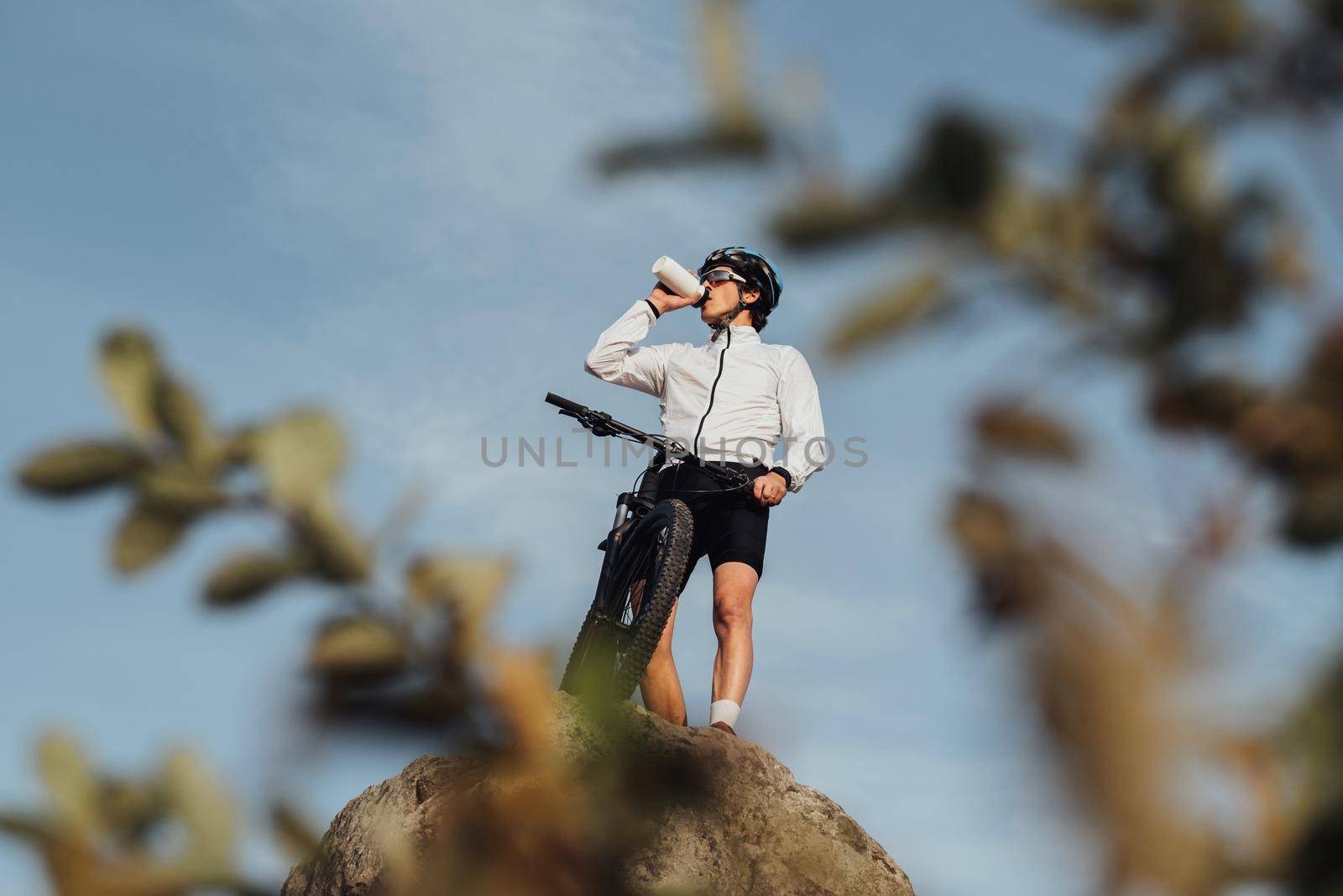 Professional Male Cyclist Drinking Water from Bottle while Standing with Bicycle on the Top of Hill by Romvy