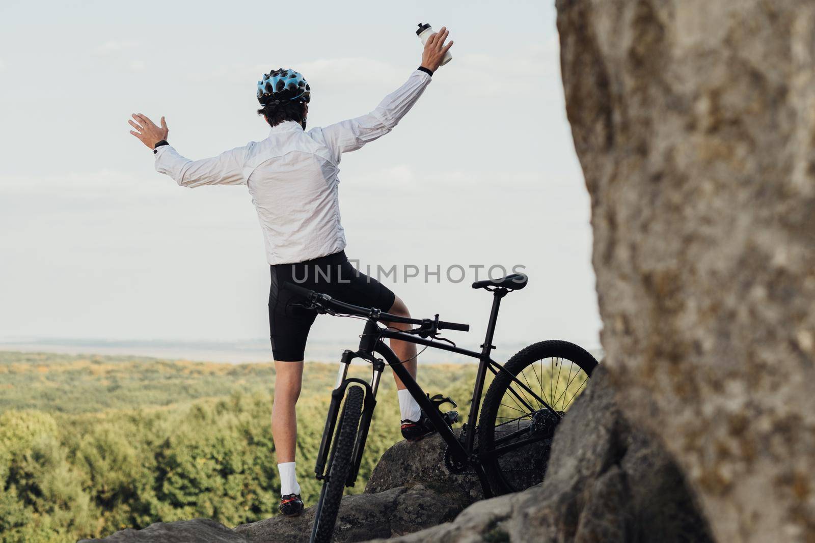 Equipped Professional Male Cyclist Sitting with Mountain Bike on Rock, Adult Man Raising Hands and Enjoying His Extreme Hobby with Panoramic View