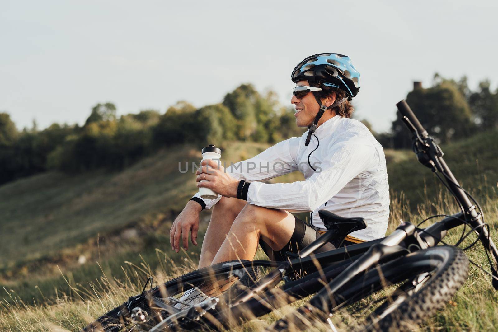 Professional Male Cyclist Drinking Water from Bottle, Man Sitting Near Bicycle During His Journey Outdoors in Countryside